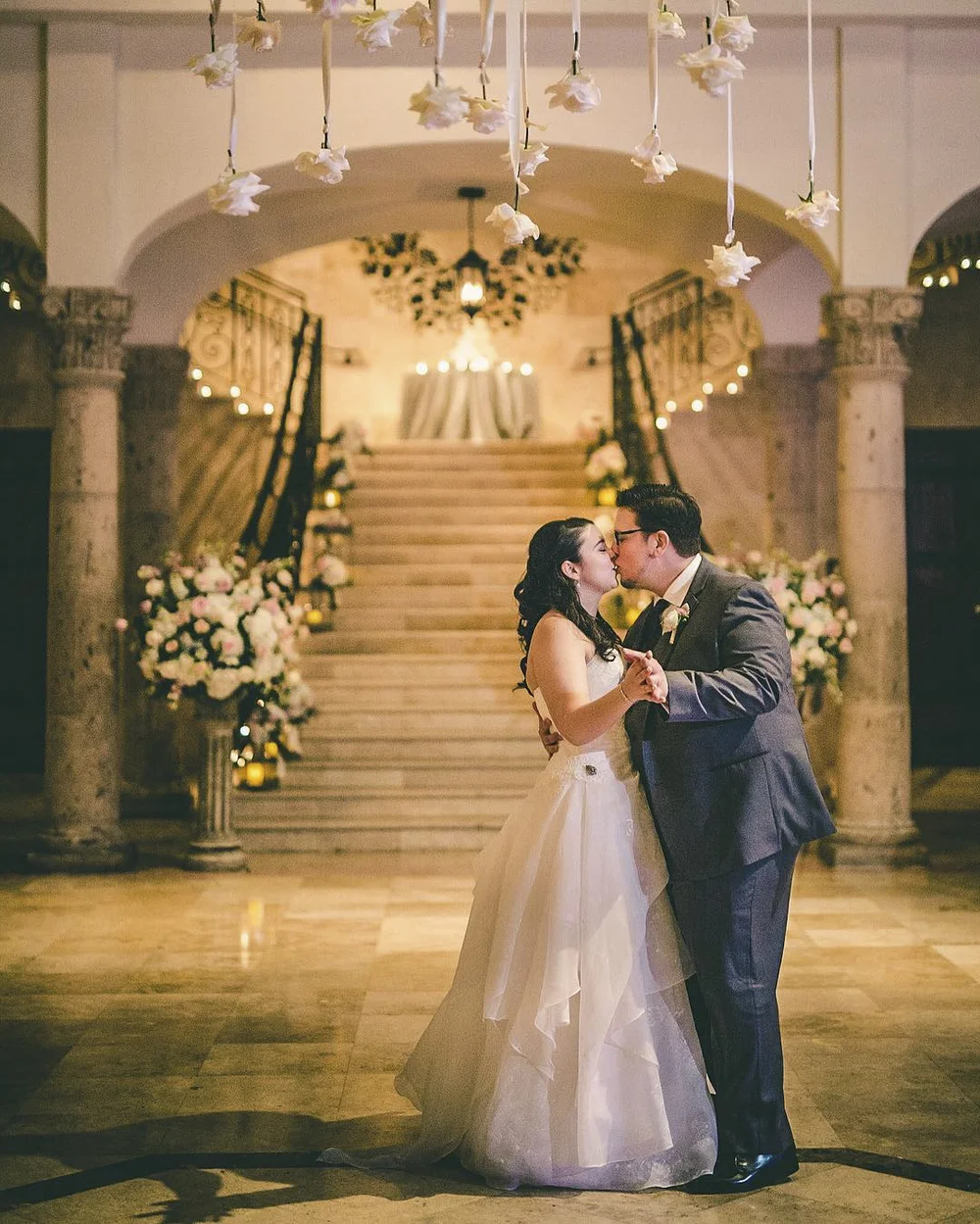 Newlywed couple dancing and kissing beneath hanging flowers with a grand staircase in the background.